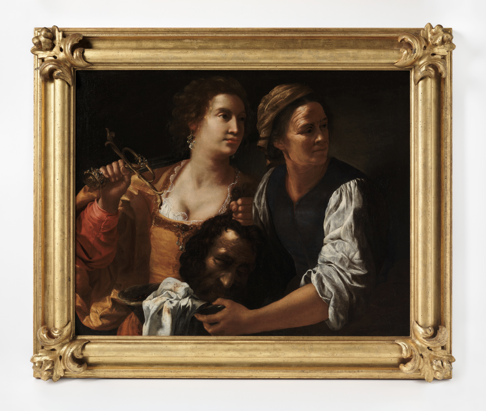 An oil painting shows two women with a light skin tone. The dark background contrasts the light complexion of the woman on the left, who is holding a sword over her shouler in one hand, and a man’s head in the other. 