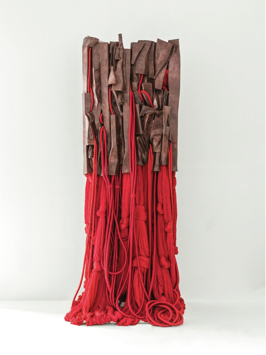 A brown and red sculpture made from leather and thick yarn stands in a room before a white wall and on a white surface. The brown leather and red yarn are knotted and intertwined with each other, creating a fluidity between the two color blocks.