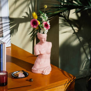 A light pink vase in the shape of a Venus, the Roman goddess of love. Her nude torso is missing both arms and she has short cropped curly hair. It sits on the corner of a wooden desk. Pink and yellow flowers sit in the vase. 
