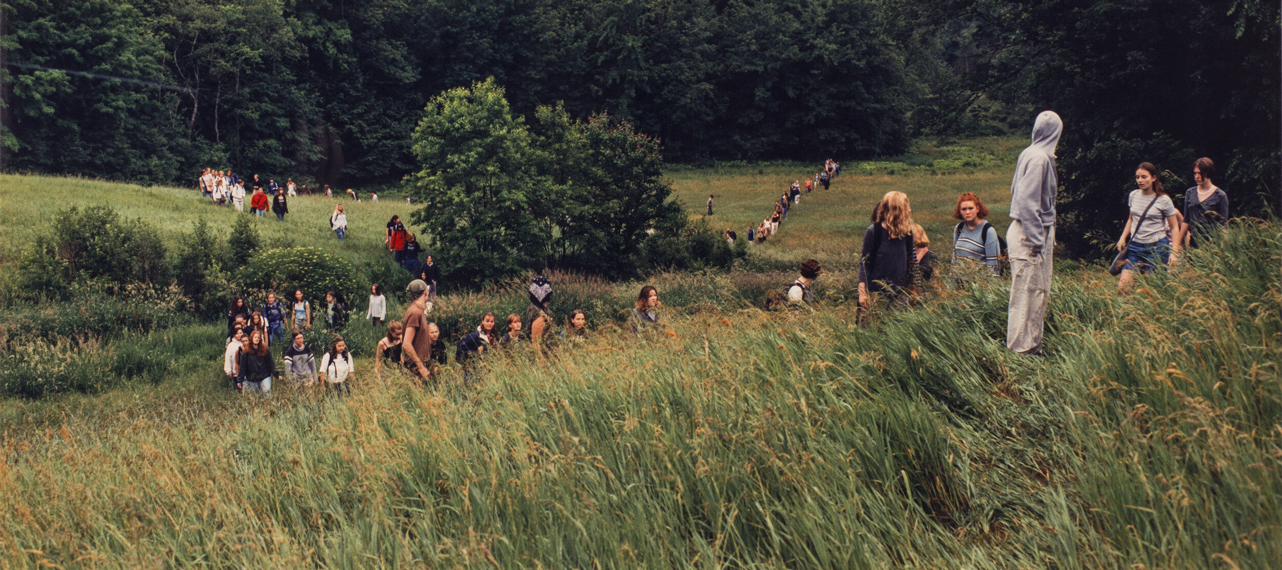 A landscape photograph featuring a field of tall green grass surrounded by leafy trees and a grey sky. A large group of people of varying ages traverse the field on foot. In the background the people form two lines, and a smaller group is congregated in the foreground.