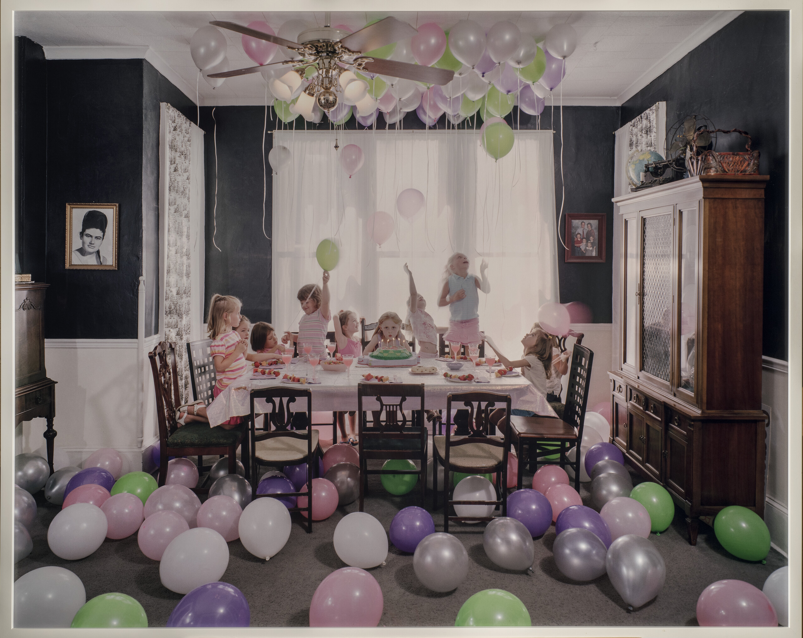 Green, purple, silver, white and pink balloons litter the floor and cover the ceiling of a navy-walled family dining room. In the middle of the room, a table is set with a pink table cloth. In the center of the table, a young girl blows out the candles on her birthday cake while looking straight ahead. Around her, nine other young girls jump, spin, and play.