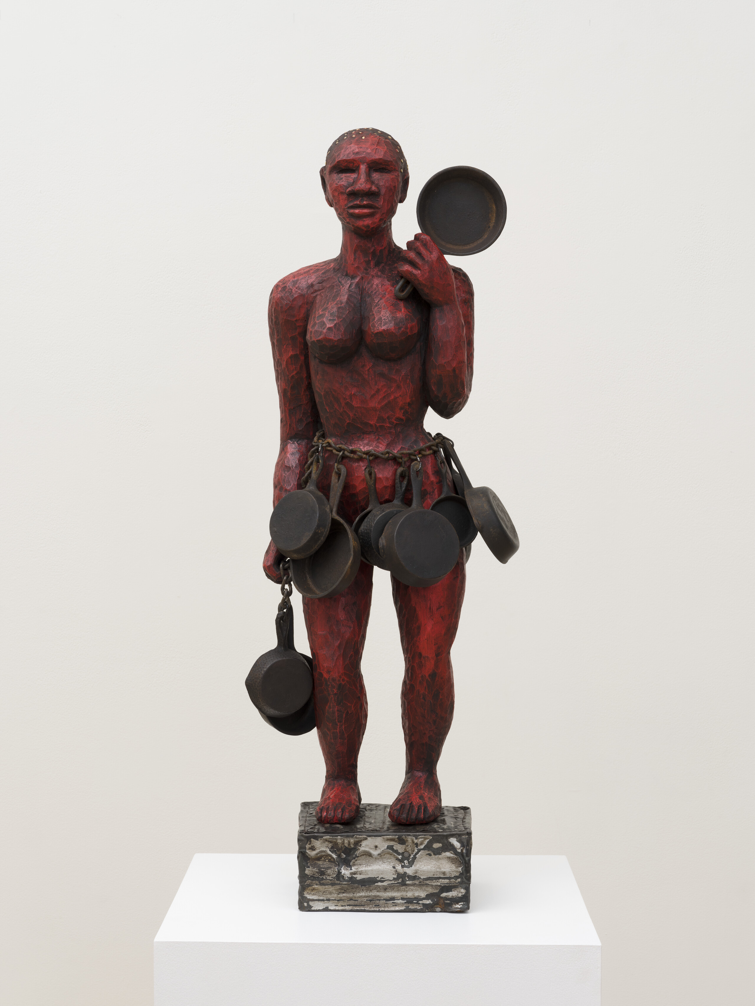 A red painted wooden sculpture of a woman holding a cast iron skillet in her left hand. She has a heavy chain around her waist with multiple skillets hanging from it like a skirt. She stands on a small wooden platform.