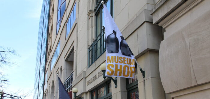 A white flag hangs on the facade of a stone building. The flag has an image of two black ceramic vases and "Museum Shop" written in gold letters below them.