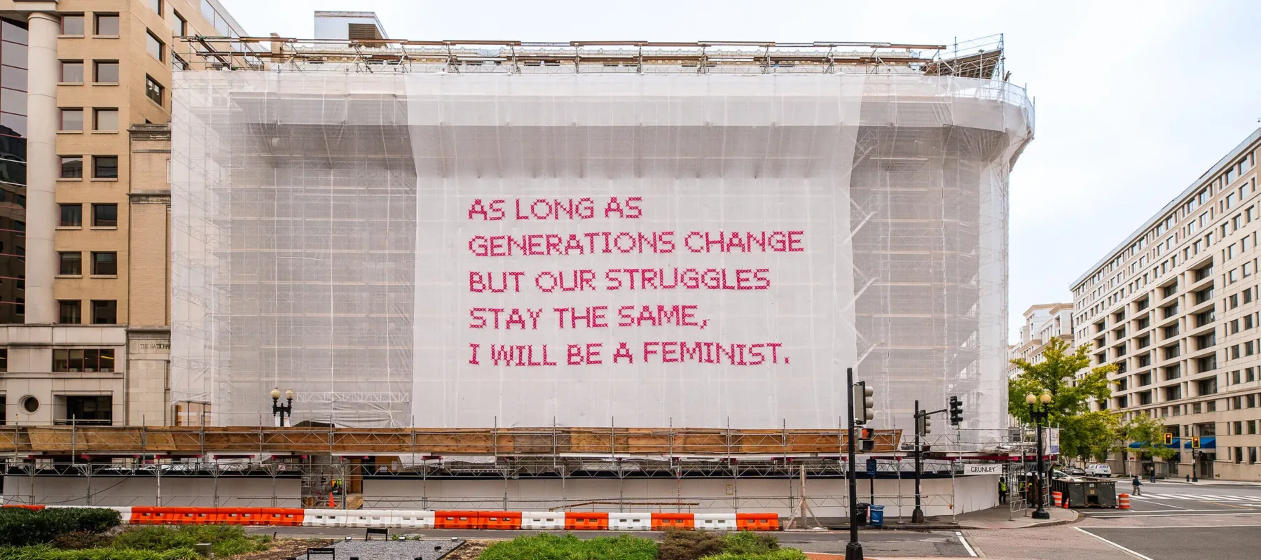 A building with a white mesh artwork covering its façade, featuring bright pink cross-stitched letters that say "As long as generations change but our struggles stay the same, I will be a feminist."