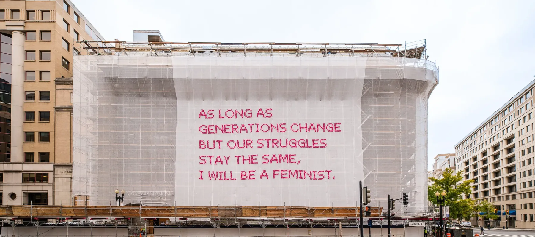 A building with a white mesh artwork covering its façade, featuring bright pink cross-stitched letters that say "As long as generations change but our struggles stay the same, I will be a feminist."