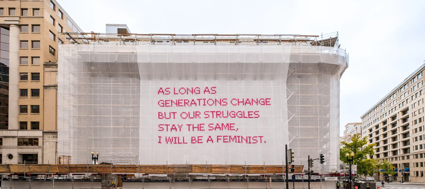 A building with a white mesh artwork covering its façade, featuring bright pink cross-stitched letters that say "As long as generations change but our struggles stay the same, I will be a feminist."