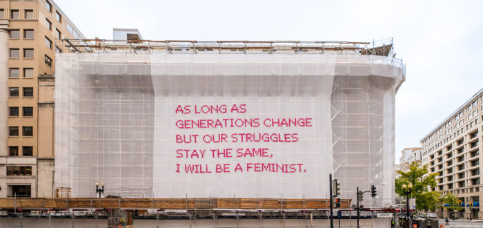 A building with a white mesh artwork covering its façade, featuring bright pink cross-stitched letters that say "As long as generations change but our struggles stay the same, I will be a feminist."