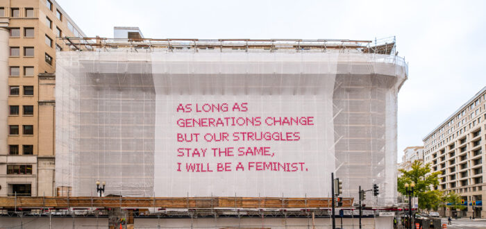 A building with a white mesh artwork covering its façade, featuring bright pink cross-stitched letters that say "As long as generations change but our struggles stay the same, I will be a feminist."