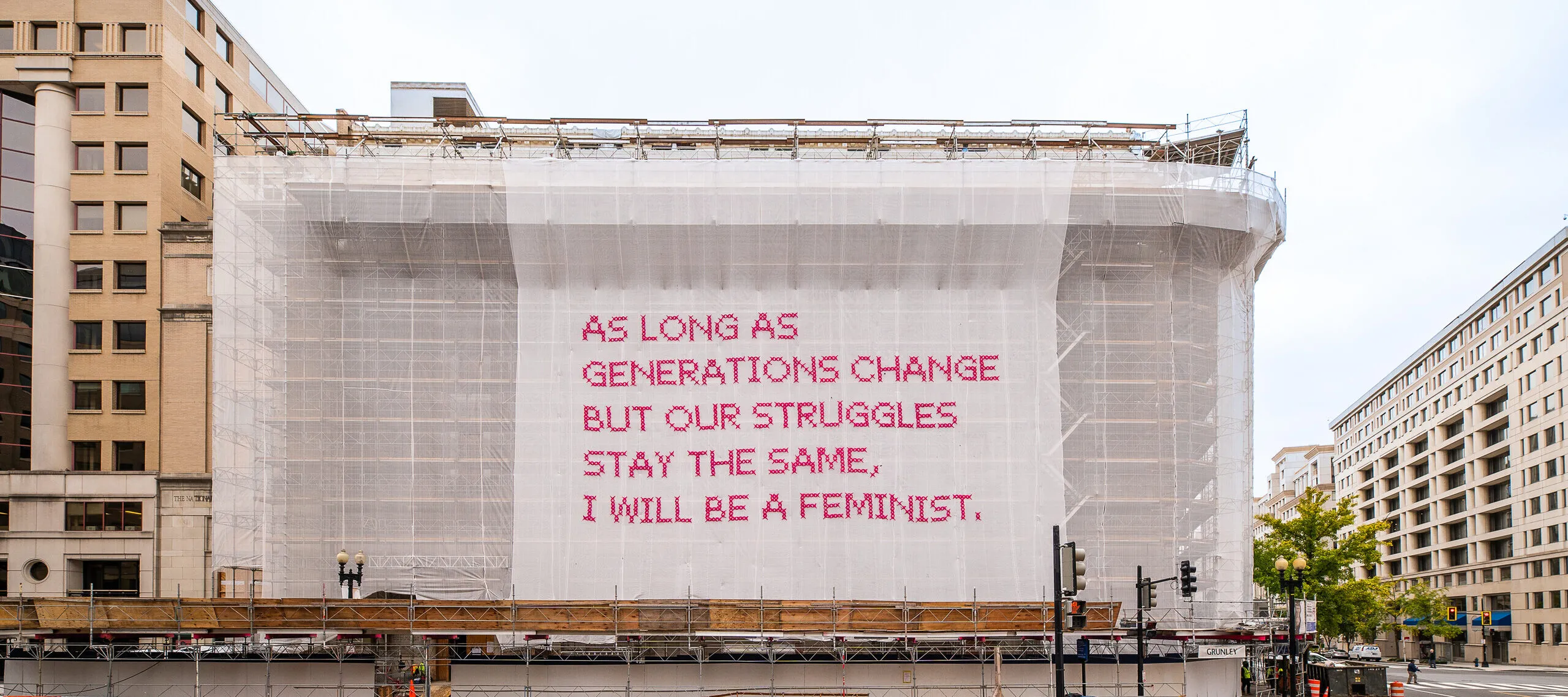 A building with a white mesh artwork covering its façade, featuring bright pink cross-stitched letters that say "As long as generations change but our struggles stay the same, I will be a feminist."