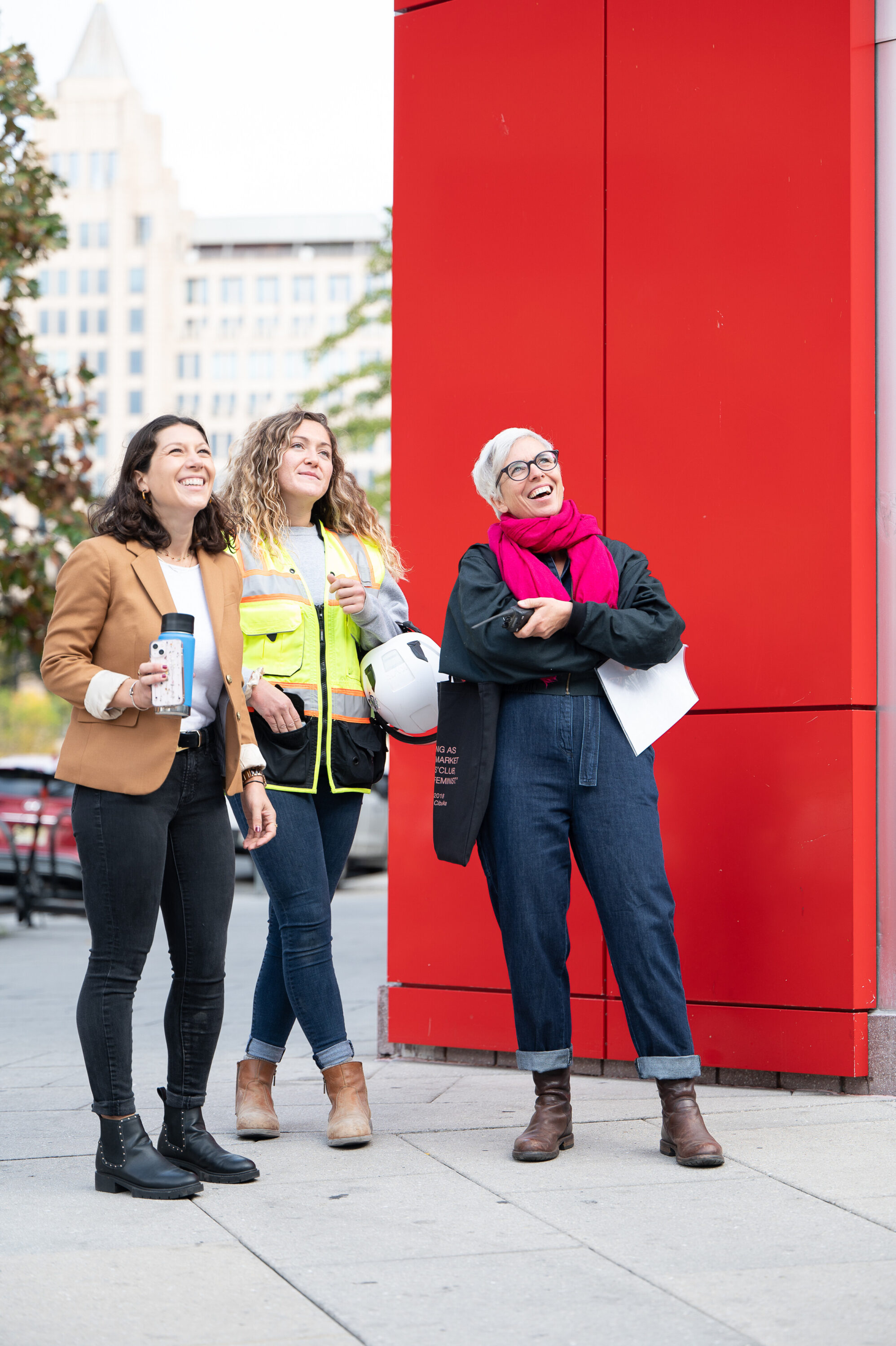 Three women with light skin tones are standing next to each other, looking up. The woman in the middle is wearing a yellow security jacket and hard hat. The woman on the right has short, gray hair and is wearing a denim overall and a pink scarf.