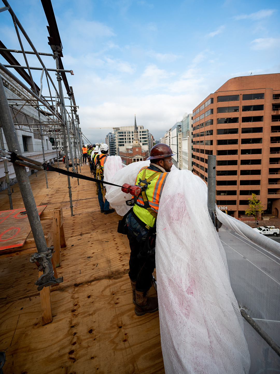 A row of people in construction hats and vests stand atop a building holding a roll of white mesh.