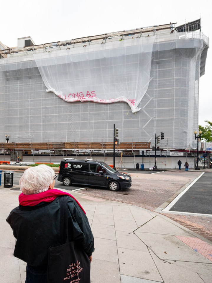 A light-skinned woman with short white hair stands in front of a building as a white mesh artwork drops down over the façade of a building.