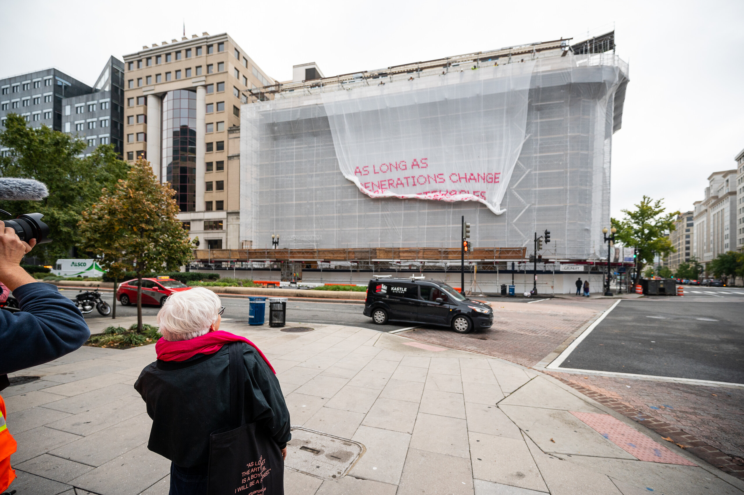 A light-skinned woman with short white hair stands in front of a building as a white mesh artwork drops down over the façade of a building, revealing bright pink text.