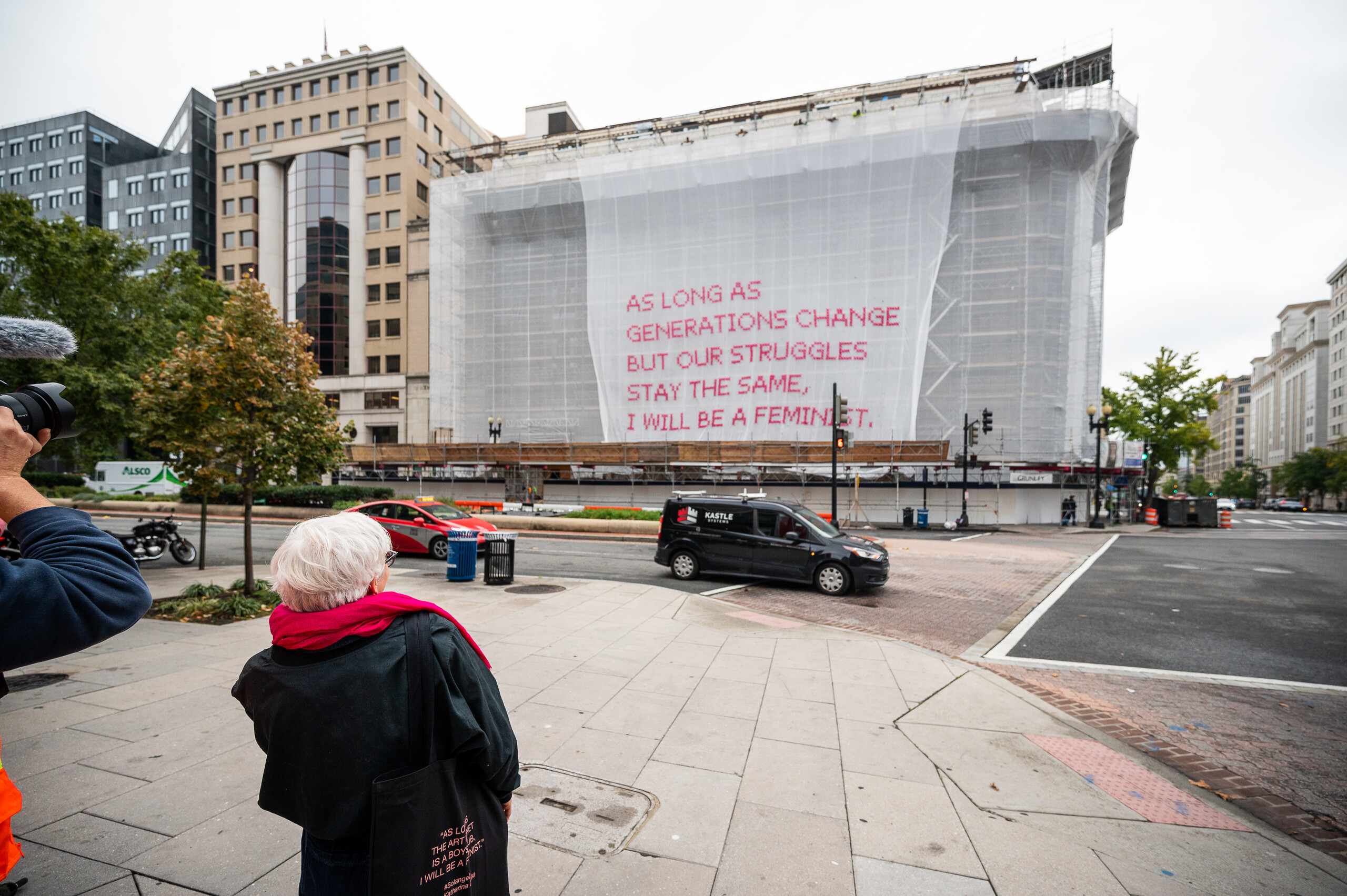 A light-skinned woman with short white hair stands in front of a building as a white mesh artwork drops down over the façade of the building, revealing bright pink cross-stitched letters that say "As long as generations change but our struggles stay the same, I will be a feminist."