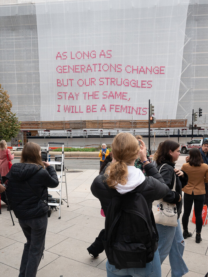 A group of people gather on a sidewalk, looking at a building covered in white mesh with the words “As long as generations change but our struggles stay the same, I will be a feminist.”