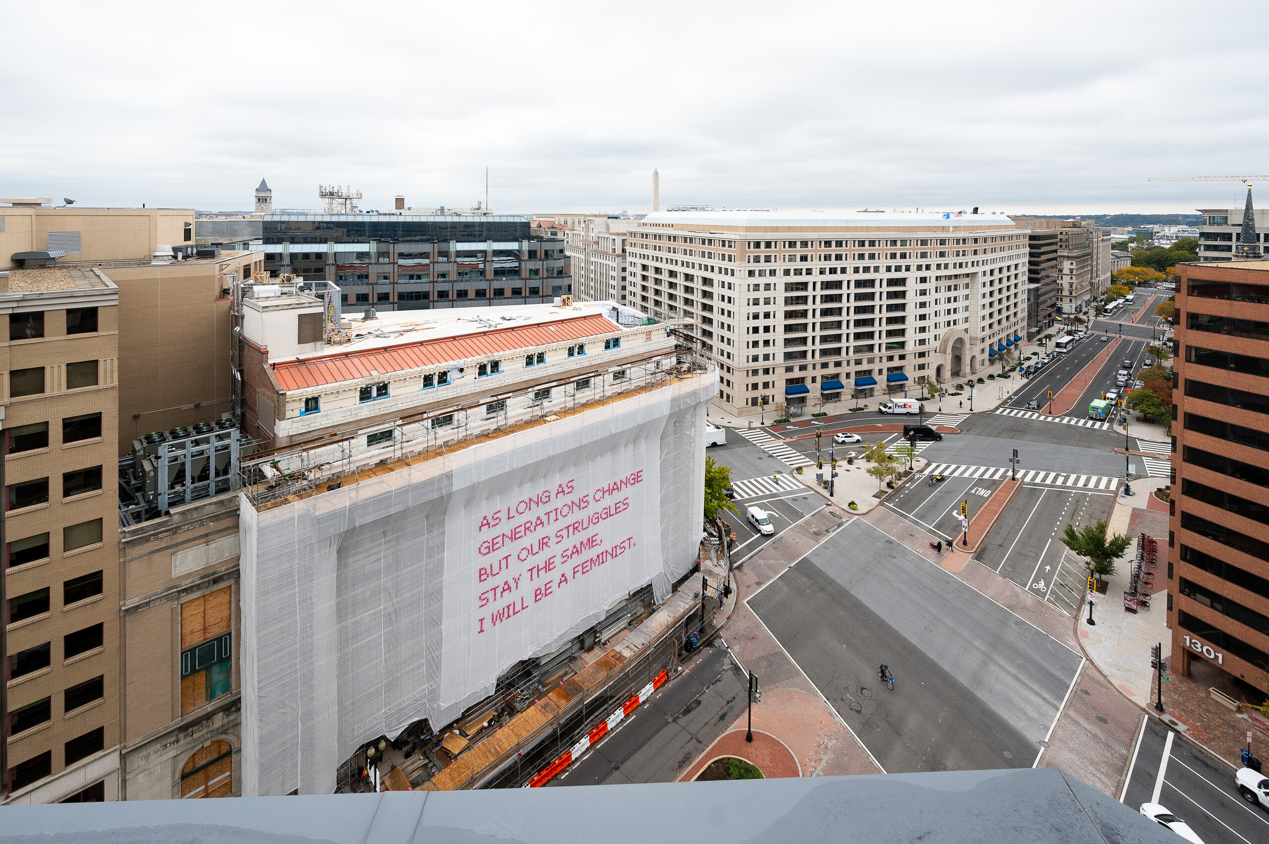 A building with a white mesh artwork covering its façade, featuring bright pink cross-stitched letters that say "As long as generations change but our struggles stay the same, I will be a feminist."