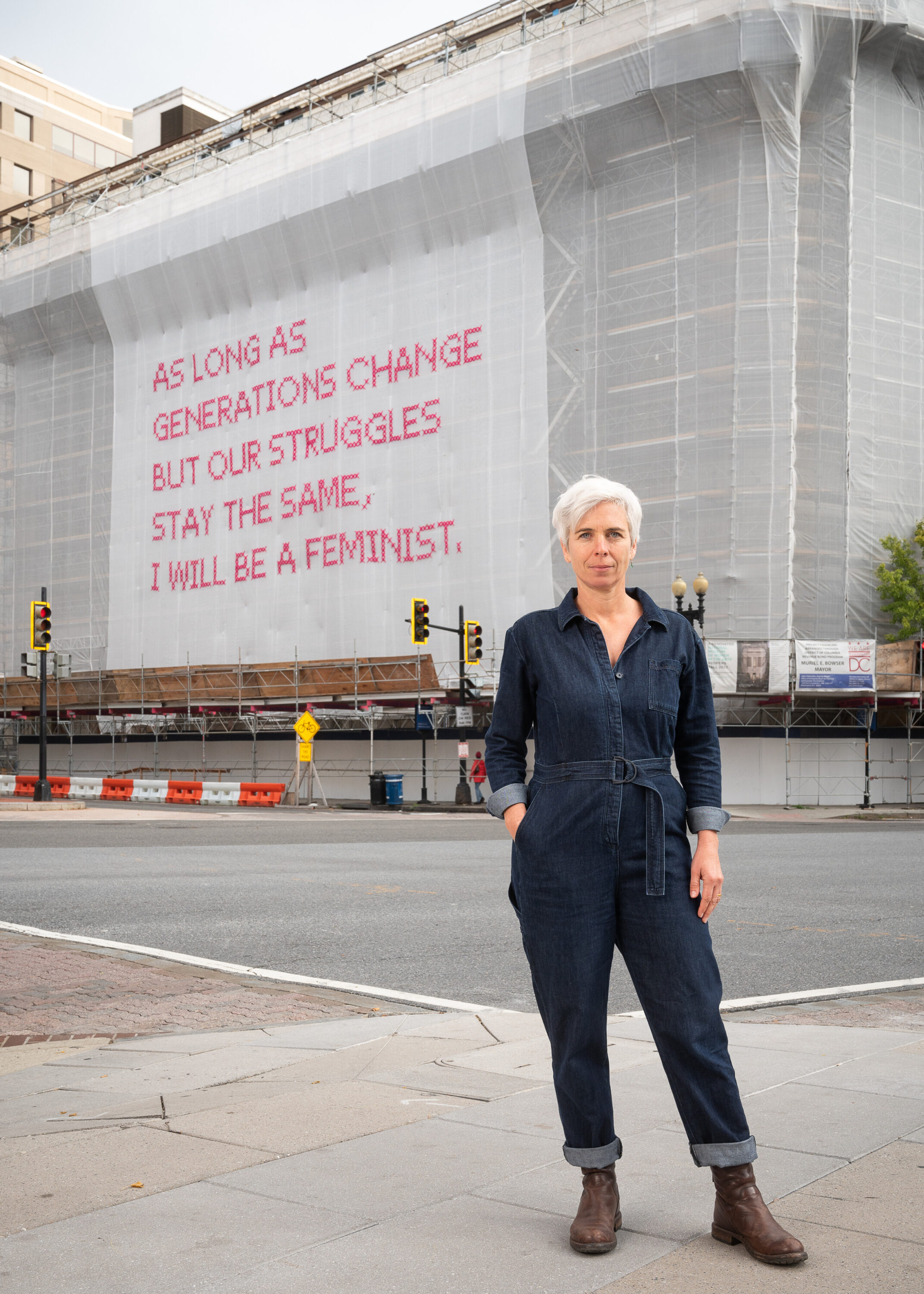 A light-skinned woman with short white hair stands in front of a building whose façade is covered in a white mesh artwork with bright pink cross-stitched letters that say "As long as generations change but our struggles stay the same, I will be a feminist."
