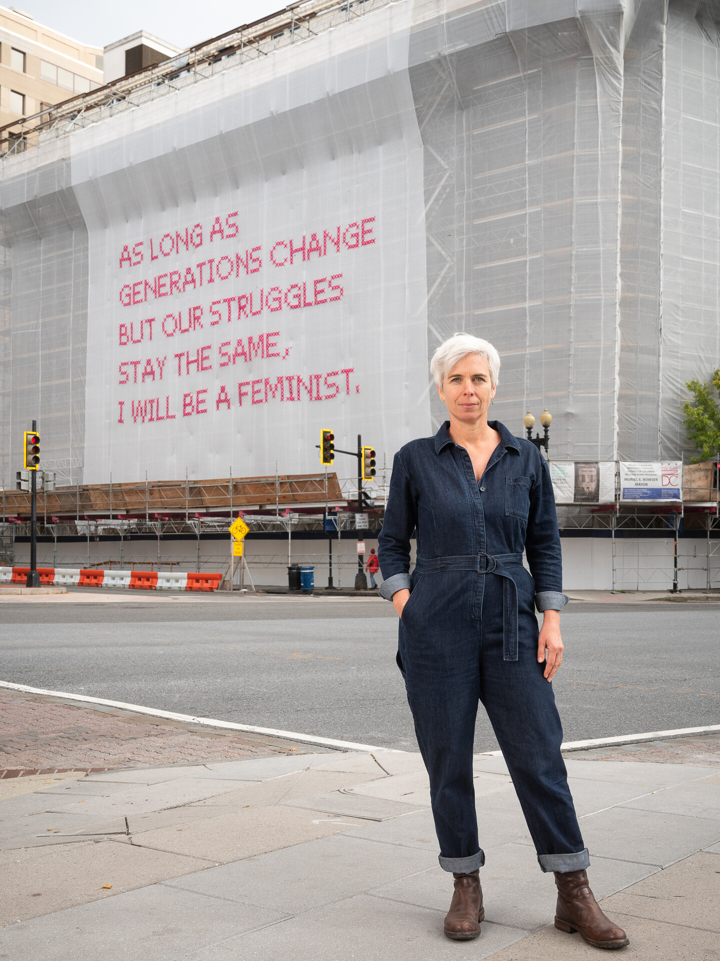 A light-skinned woman with short white hair stands in front of a building whose façade is covered in a white mesh artwork with bright pink cross-stitched letters that say 