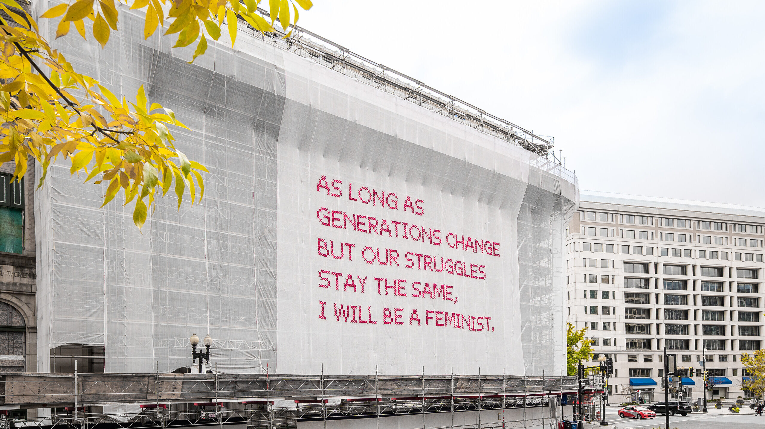 A building with a white mesh artwork covering its façade, featuring bright pink cross-stitched letters that say 