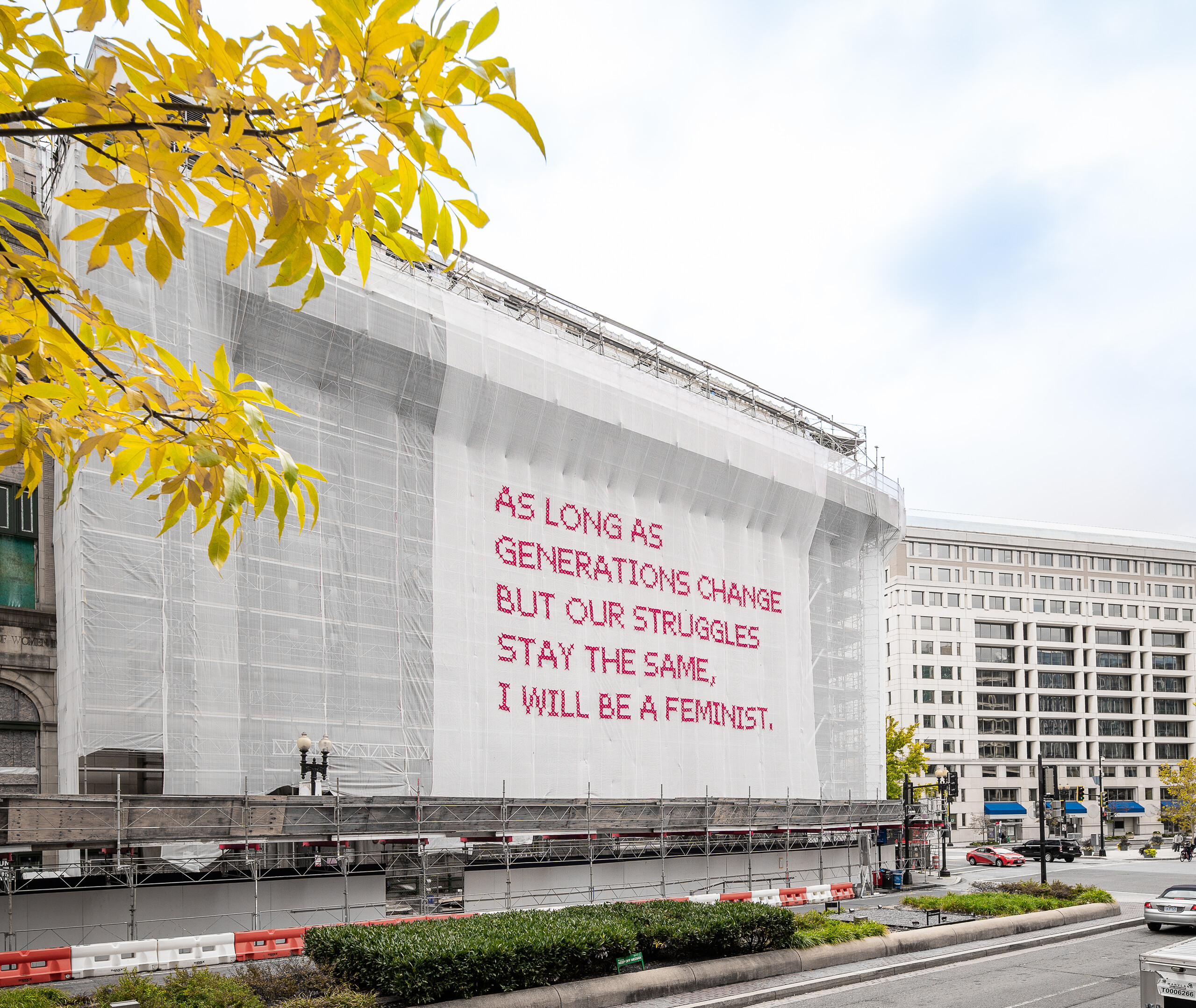 A building with a white mesh artwork covering its façade, featuring bright pink cross-stitched letters that say "As long as generations change but our struggles stay the same, I will be a feminist."