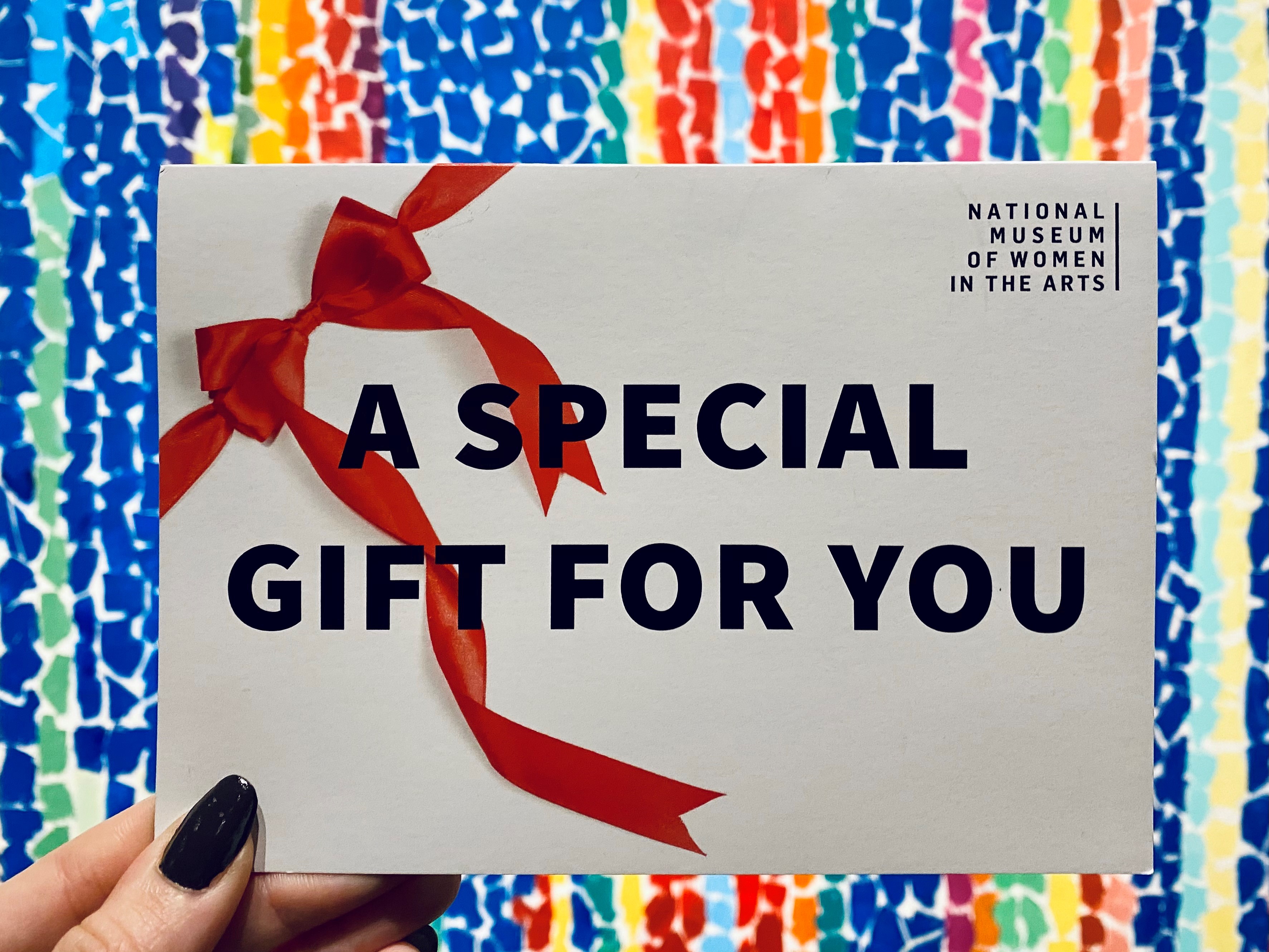 A light-skinned hand holds an envelope tied with a red ribbon that reads “A Special Gift for You” with the logo for the National Museum of Women in the Arts in the upper right corner. Behind the envelope is a colorful abstract painting.