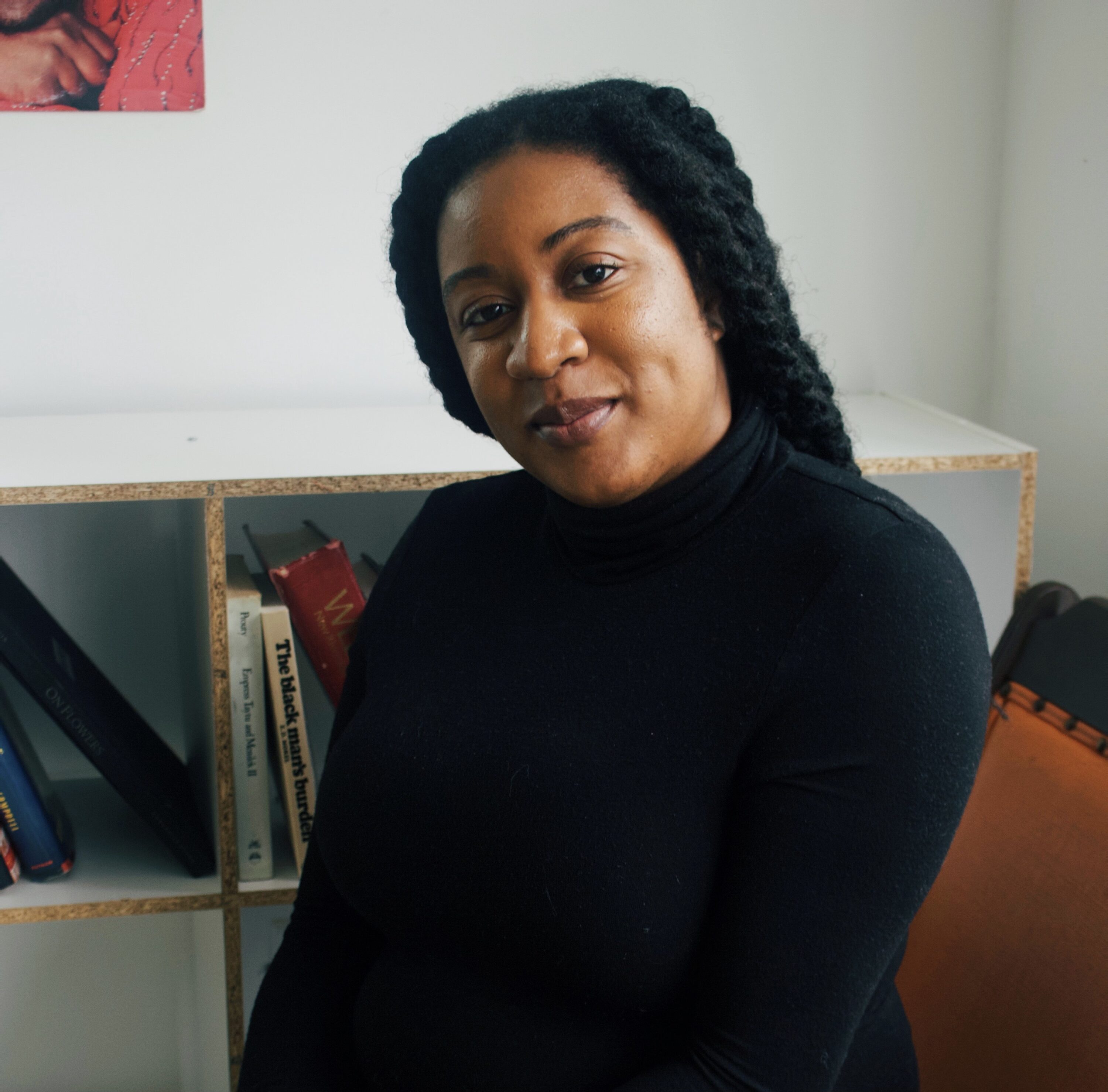 A woman with a medium-dark skin tone sits before a white shelf stacked with books and smiles for a portrait photo. The woman has long, black hair and wears a black turtleneck; natural light illuminates her face from the side. 