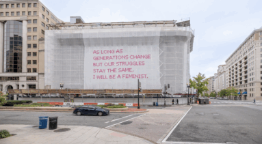 Video still of a building with a white mesh artwork covering its façade, featuring bright pink cross-stitched letters that say "As long as generations change but our struggles stay the same, I will be a feminist."