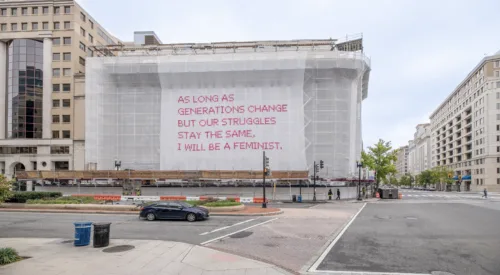 Video still of a building with a white mesh artwork covering its façade, featuring bright pink cross-stitched letters that say "As long as generations change but our struggles stay the same, I will be a feminist."