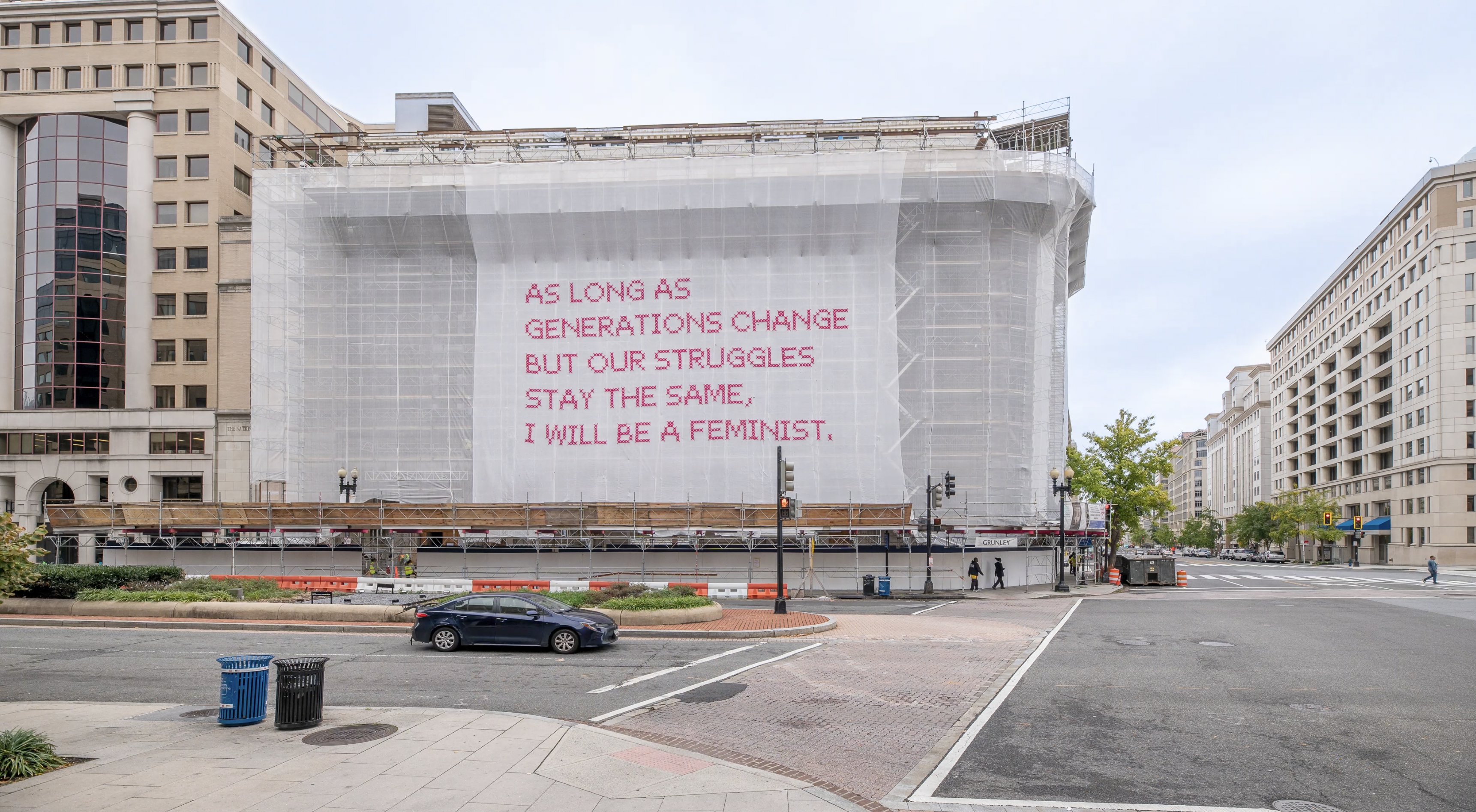 Video still of a building with a white mesh artwork covering its façade, featuring bright pink cross-stitched letters that say 
