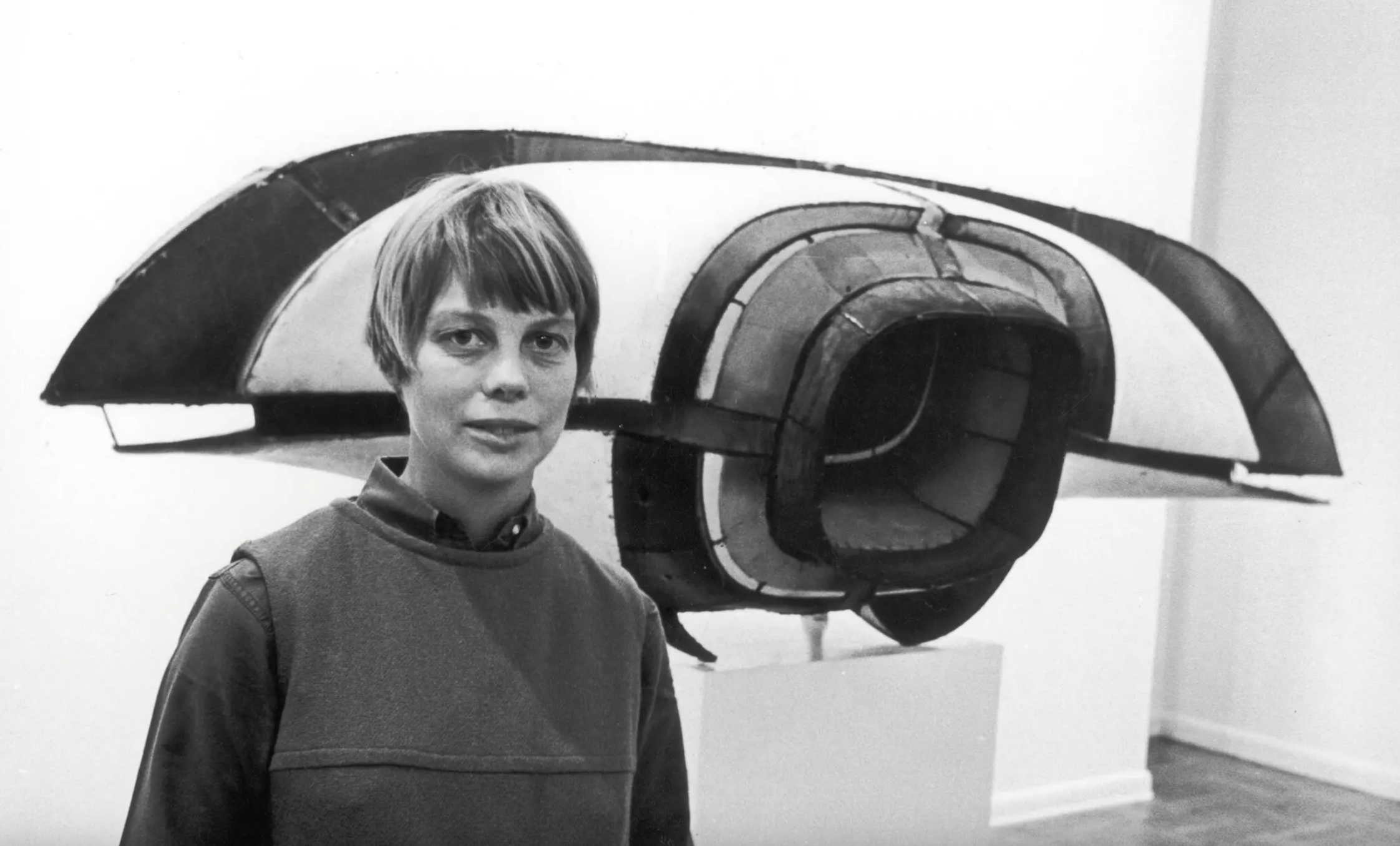 A black-and-white photograph of a woman with a light skin tone standing in front of a sculpture. The woman has blonde, short hair and looks directly into the camera. The sculpture is made from metal and resembles parts of an airplane.