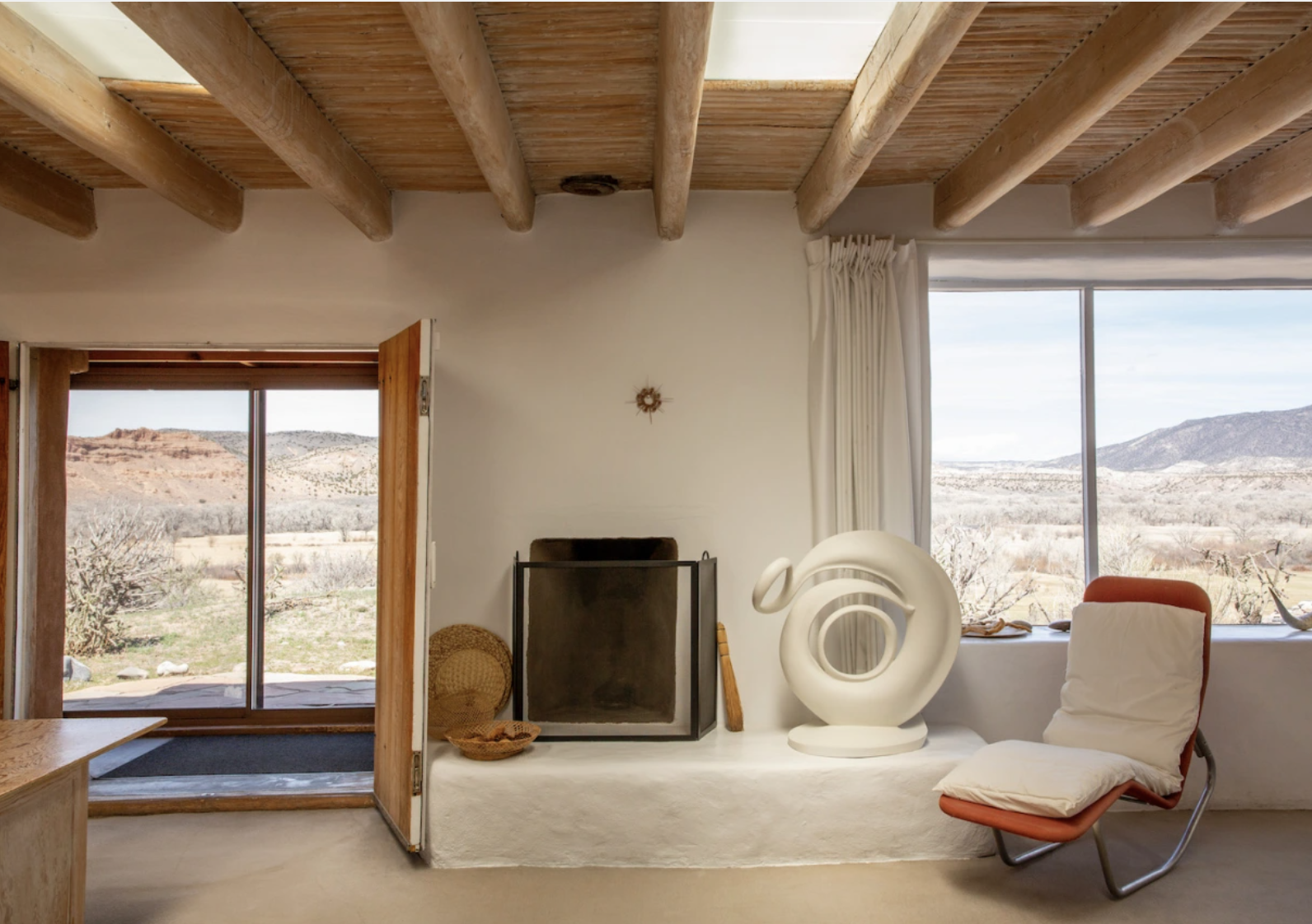 A photograph shows an interior of a room overlooking the New Mexican desert. The room is in beige tones and has a wood ceiling. Through big windows, plains and a mountain are visible in the distance. The color palette inside resembles the colors outside.