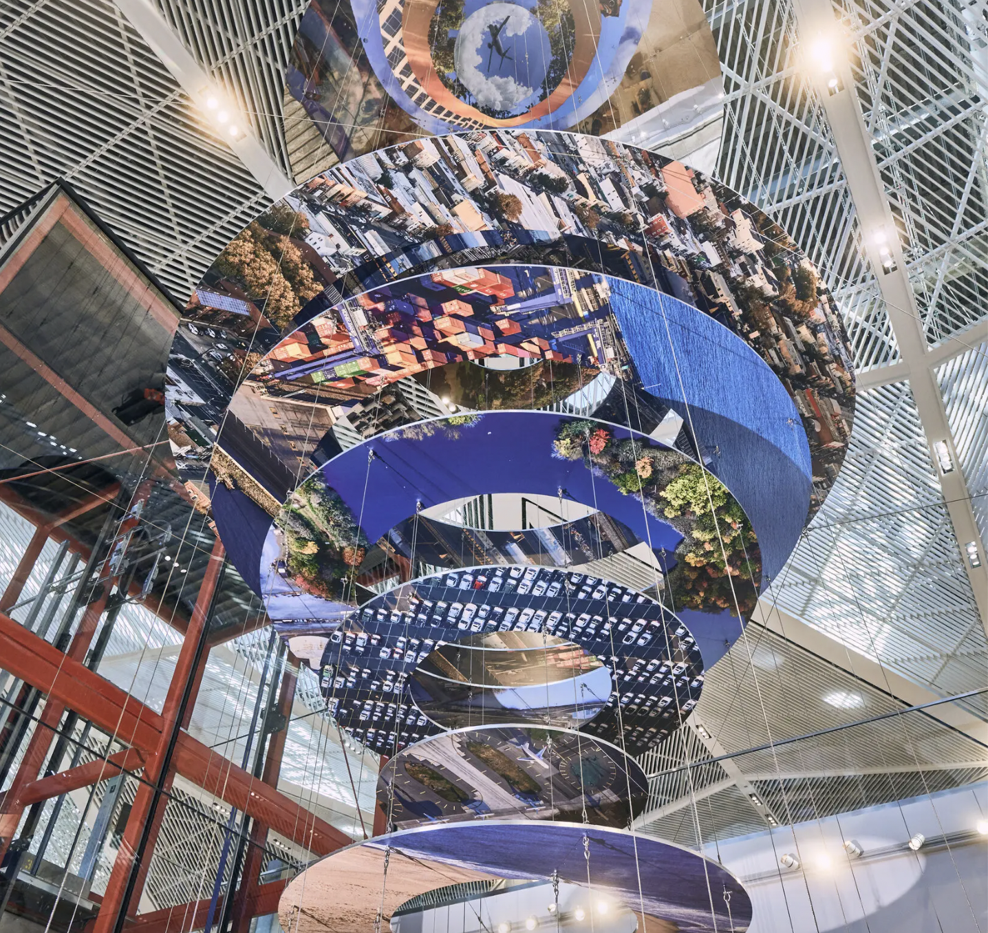 A series of disks hangs from the ceiling of a large, industrial building. The disks are covered in photographs shot from the aerial perspective, including a car park, a beach, city buildings, and a harbor freight transport. The sculpture as a whole seems very futuristic, evoking notions of satellites observing earth.