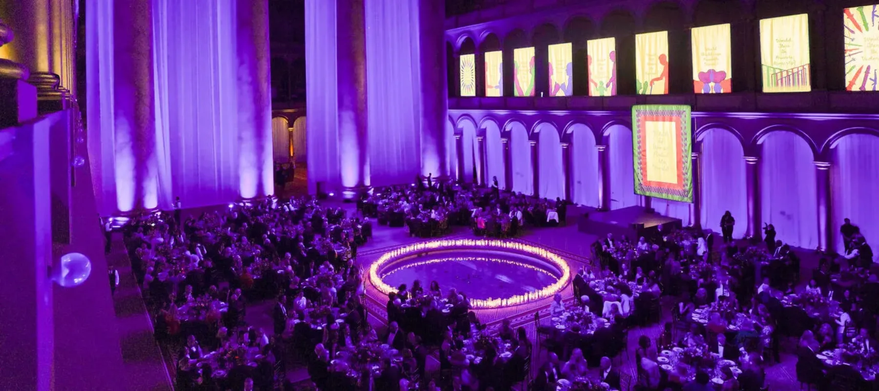 Interior of a large room filled with people sitting around many round tables. There is a circle of candles in the center of the room, and purple lighting fills the space. In the background, there are four massive columns and ten panels of artwork hanging on the adjacent wall.