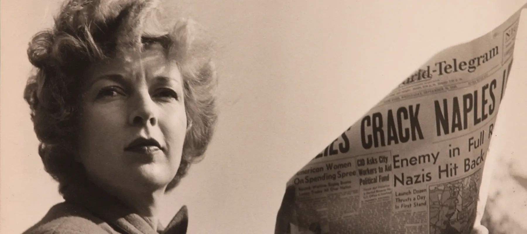 A black-and-white photograph of a light-skinned adult woman holding a newspaper with news about World War II. She wears a coat and her short, curly hair is caught in the wind.