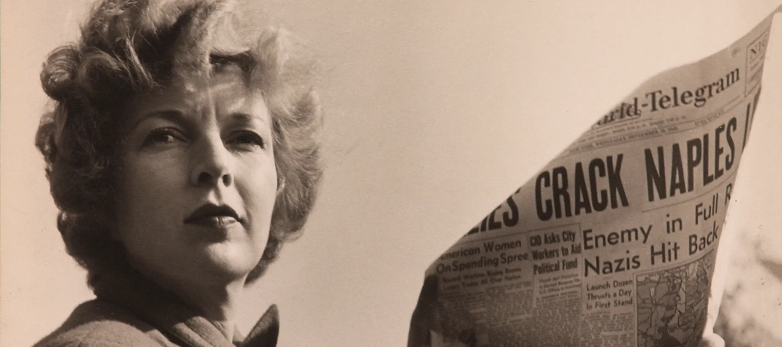 A black-and-white photograph of a light-skinned adult woman holding a newspaper with news about World War II. She wears a coat and her short, curly hair is caught in the wind.