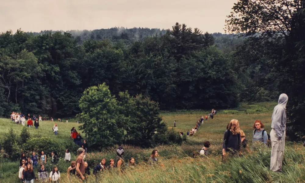 A landscape photograph featuring a field of tall green grass surrounded by leafy trees and a grey sky. A large group of people of varying ages traverse the field on foot. In the background the people form two lines, and a smaller group is congregated in the foreground.