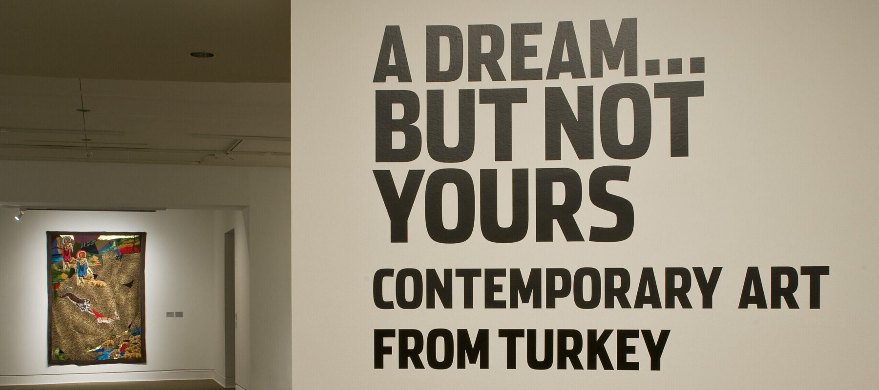 Installation view of a gallery space. On a white wall, it says in big black letters "A Dream... but not yours. Contemporary Art from Turkey." In the back, there is a large painting hanging on another wall.