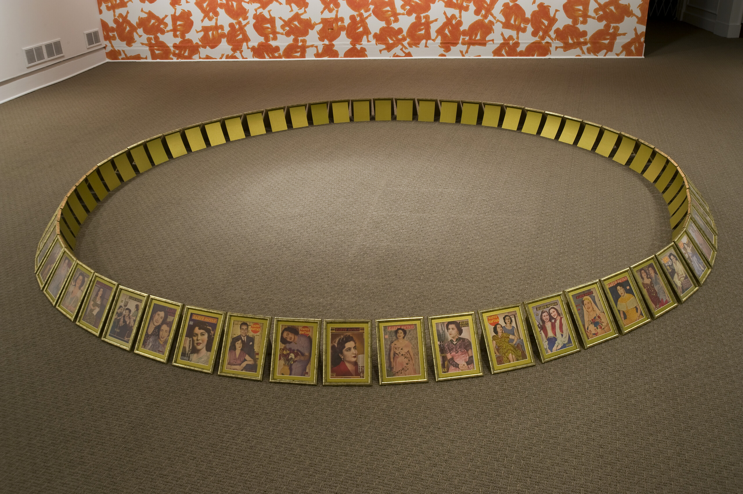 On a gray carpet floor, framed images of women in golden frames are placed in a large circle. The images of the women are vintage-looking. On the wall behind the circle, a large flower print is installed, that looks like a wallpaper, taking up the whole wall. The images in the golden frames look precious, and their placement in a circle creates a community of women.