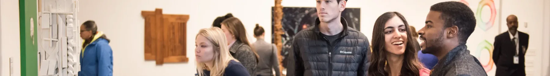 Several museum visitors look at various paintings and sculptures throughout a gallery while a security guard in the background monitors.