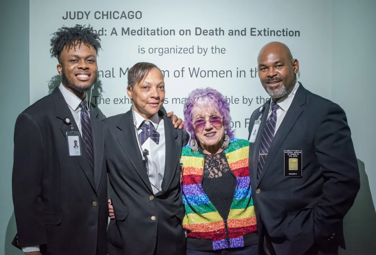 Three Security Officers and artist Judy Chicago stand next to each other smiling in front of opening text to the artist's exhibition.