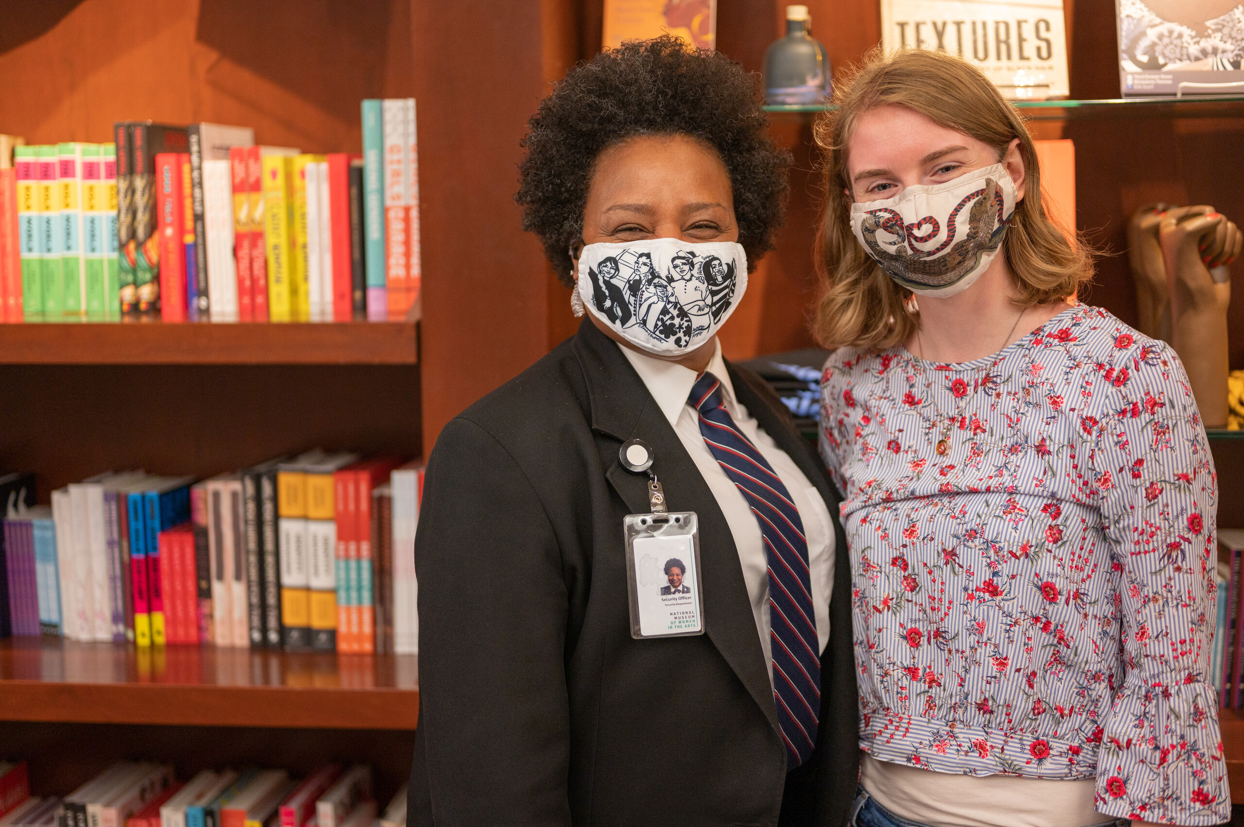 A dark-skinned woman with a short afro and a light-skinned woman with strawberry-blonde shoulder-length hair pose together in front of wooden bookshelves. They both smile behind face masks featuring artsy illustrations.