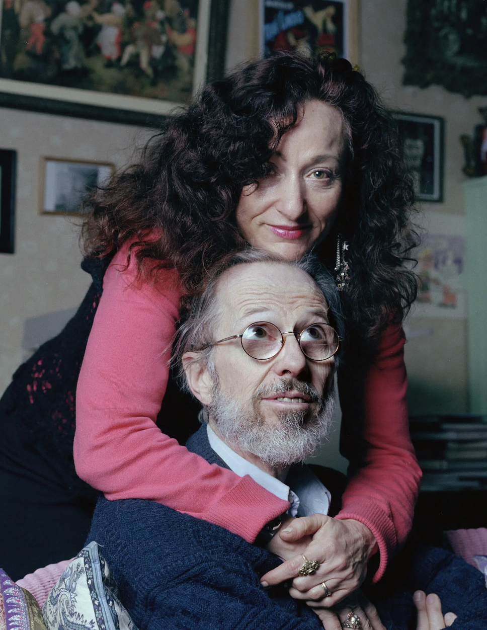  A woman with a light skin tone and long brown, curly hair wraps her arms around a man sitting in a chair and looks directly into the camera. The man has a light skin tone and a white beard, wears thick glasses, and looks up at her. She is wearing a pink shirt and pink lipstick, smiling into the camera. Behind them is a wall full of art.