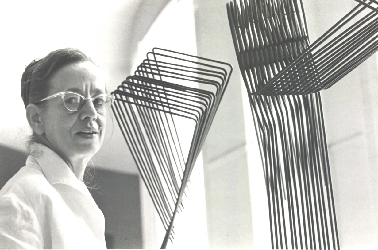 A black-and-white photo shows a woman with a light skin tone and transparent glasses standing next to a sculpture made of bent wires. The sculpture has an industrial feel due to the material and geometric form, and stands in stark contrast to the light and airy backdrop. 