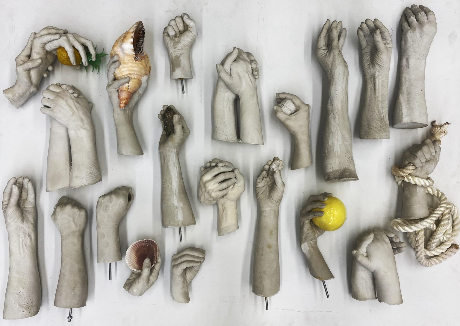 Various hands made of plaster lie on a white surface. They have a lifelike appearance indicating they are modeled after people’s hands. Each hand is holding an item: A lemon, a shell, a pineapple, and other objects. The lifelike appearance and gray tone makes them look like real, chopped off hands.
