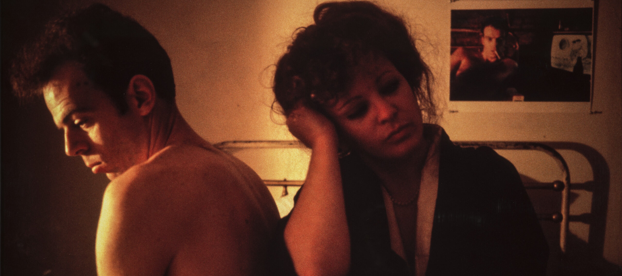 A man and woman sit next to each other on a bed. The man, topless, sits with his back to the woman. She sits leaning against him and looking away, her head resting on her hand and her elbow on her bent knee. A photo on the wall behind them appears to be of the same man.