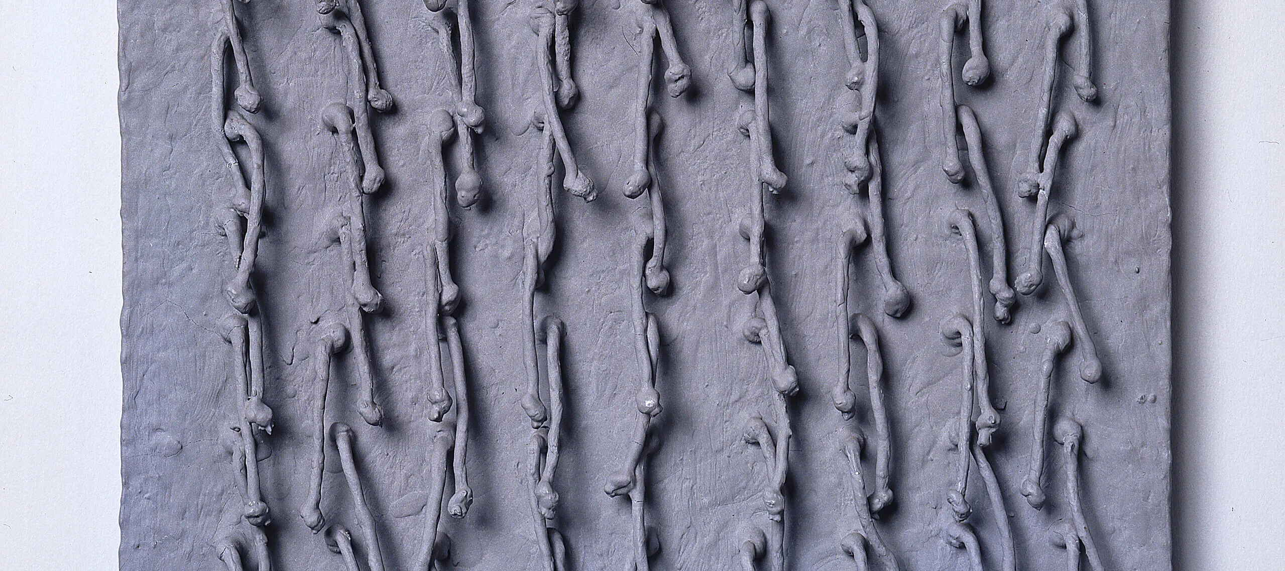 Short, knotted cords, arranged in a symmetrical 9-by-9 grid, emerge from a square panel and hang down. Thick, matte paint in a silvery gray color coats the entire piece, adding rough texture to the panel and stiffening the cords into positions that cast irregular shadows across the work’s surface.