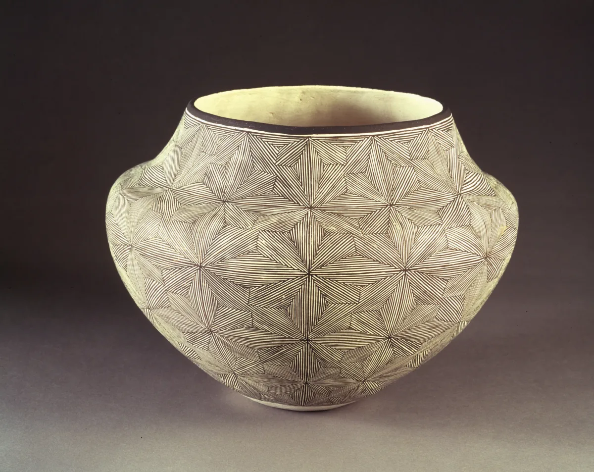 Ceramic jar, featuring a short neck and broad shoulders tapering to a narrow base, is decorated in a geometric, black and white quilt-like pattern. The matte, off-white surface is adorned with  geometric, flower-like patterns created by very thin, precisely-placed black lines.