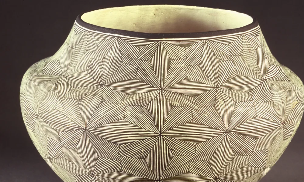 Ceramic jar, featuring a short neck and broad shoulders tapering to a narrow base, is decorated in a geometric, black and white quilt-like pattern. The matte, off-white surface is adorned with  geometric, flower-like patterns created by very thin, precisely-placed black lines.