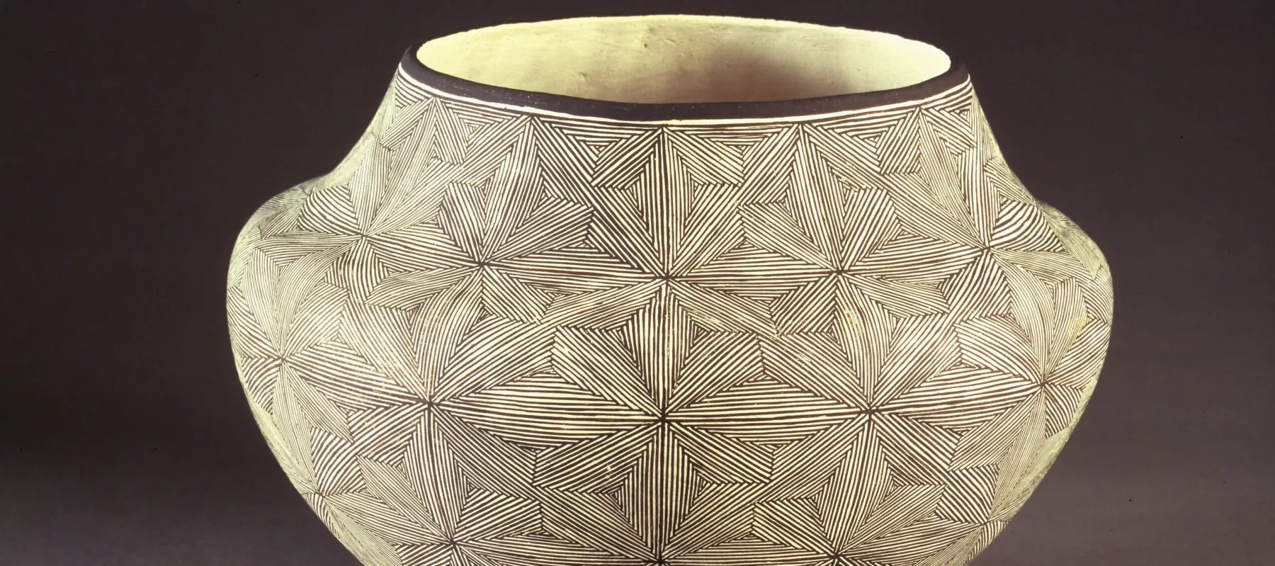 Ceramic jar, featuring a short neck and broad shoulders tapering to a narrow base, is decorated in a geometric, black and white quilt-like pattern. The matte, off-white surface is adorned with  geometric, flower-like patterns created by very thin, precisely-placed black lines.