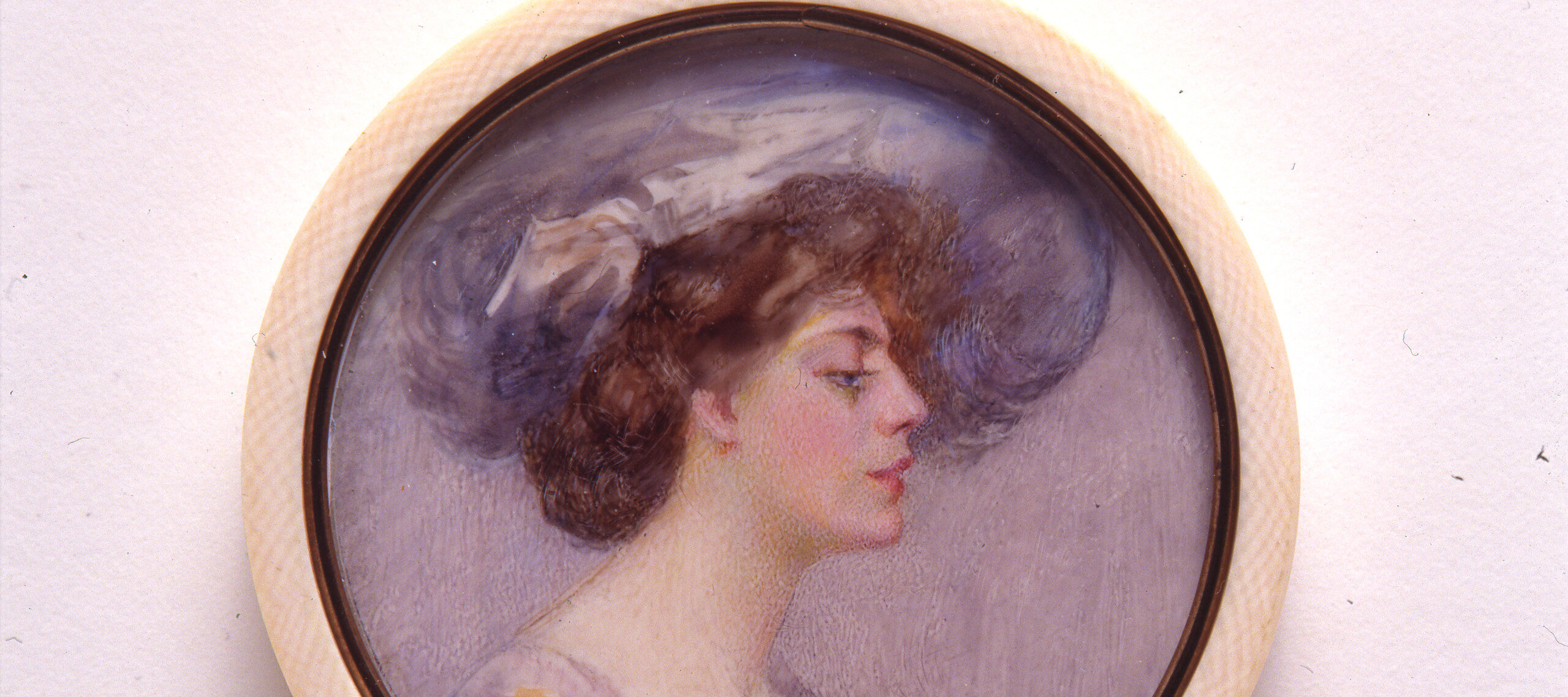 A miniature portrait in a round white frame of a light skinned woman in profile with brown hair pulled back in a loose bun. The woman wears a large periwinkle hat that matches the sleeves of her purple and blue dress. The background of the portrait is pale purple.